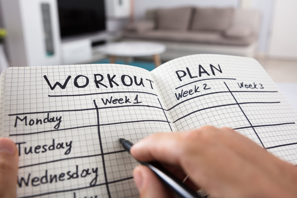 A hand holding a pen writing a fitness plan on a 5 day workout schedule.